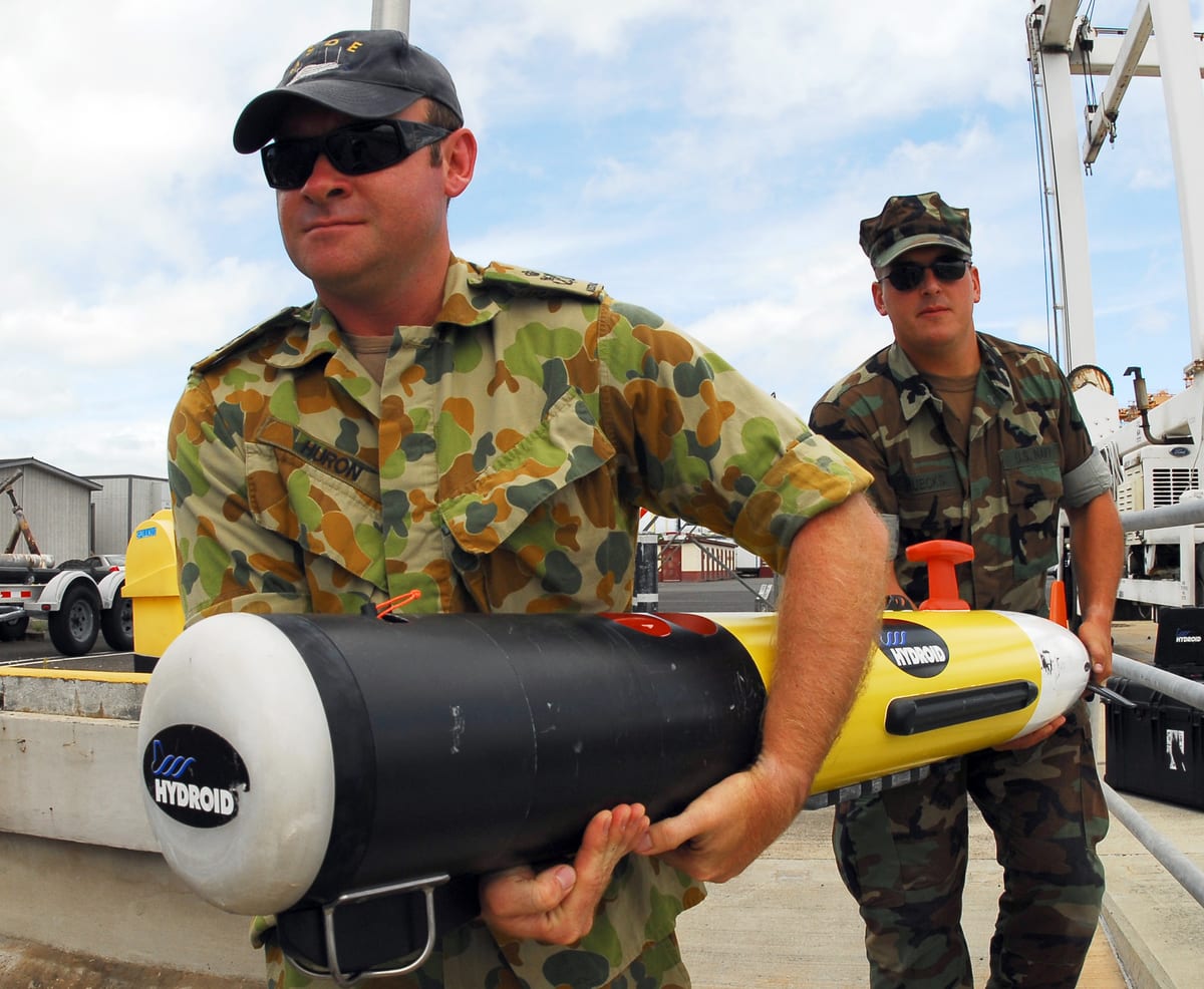 U.S. and Australian Navy personnel carrying a yellow REMUS 100 autonomous underwater vehicle
